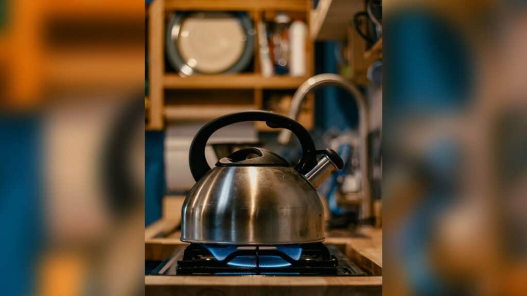 A kettle heating up on a gas burner. In the background there is a plate and paper towels hanging on the wall. the background is slightly blurred out while the foreground of the kettle is very clear.