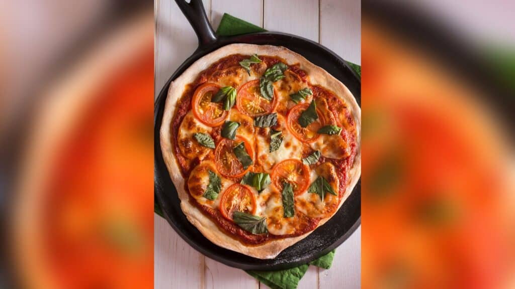 Picture is off a pizza that was baked in cast iron pan. It is a top down image with the handle of the cast iron pan pointing to the upper left of the image. The pan is sitting on a wood table.