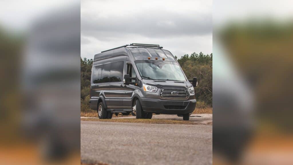 2015 Ford Transit in gray. The van has green trees behind it and a cloudy sky