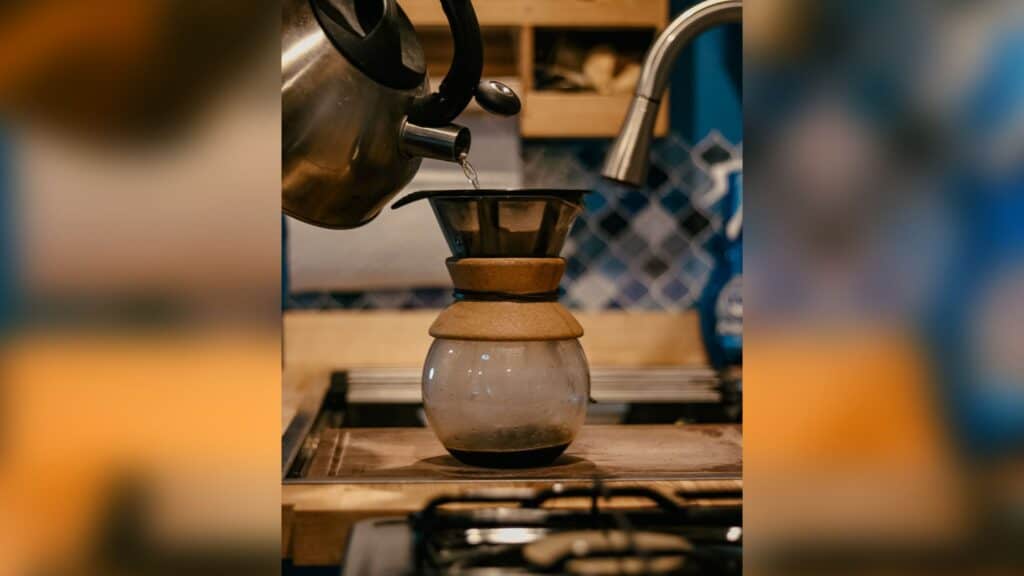 A kettle is pouring water into the Bodum pour over. The pour over is sitting on a cutting board. The pour over has a little bit of coffee in the bottom. The background is burred out, but you can see blue tiles in the back.