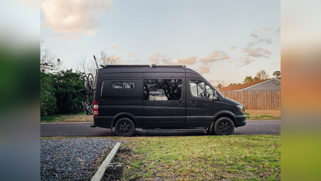 An all black Sprinter van that is converted is parked on a street. There is grass leading up the the van, and an orang wood fence behind the van in the background