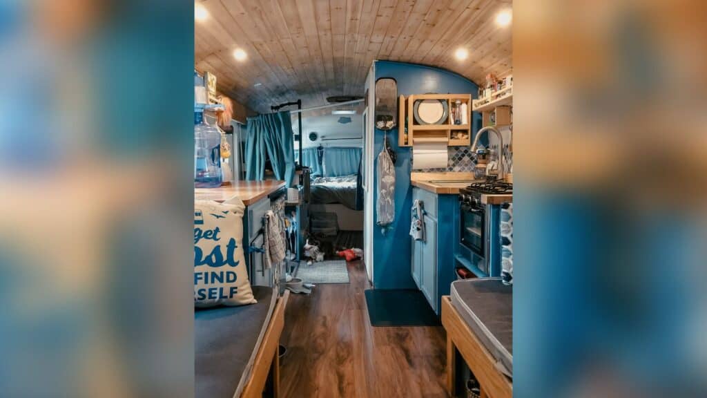 The inside of a bus conversion. There are dark wood floors, a cedar plank roof, and a kitchen in view. The kitchen is painted blue and have raw wood butcherblock countertops