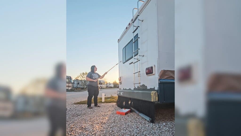 The photo is showing how to paint an RV. The photo is of the back of an RV with a person using a paint roller on an extension stick to paint