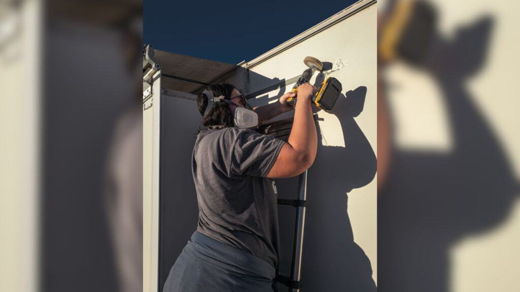 Showing how to paint an RV, There is a person center frame using a 3M eraser wheel attached to a power drill removing decals. 